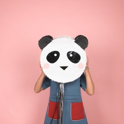 Piñata com rosto de panda segurada por pessoa com vestido azul e bolsos vermelhos, fundo rosa