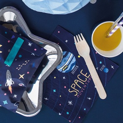 Guardanapo e embalagem de tecido azul com padrão espacial, prato prateado estrela, copo branco com sumo laranja e palhinhas e garfo de madeira