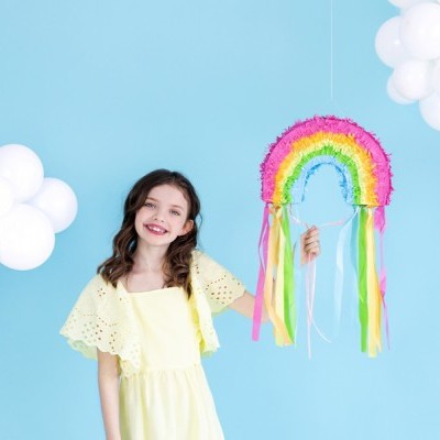 Menina sorridente com vestido amarelo segurando piñata de arco-íris colorido com franjas e fitas, fundo azul e balões brancos em forma de nuvens