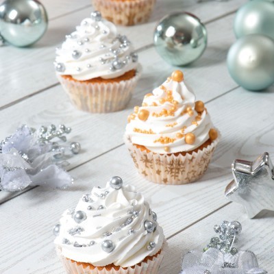 Três cupcakes decorados com cobertura branca e pérolas comestíveis em cima, no meio de enfeites prateados e bolas de Natal.