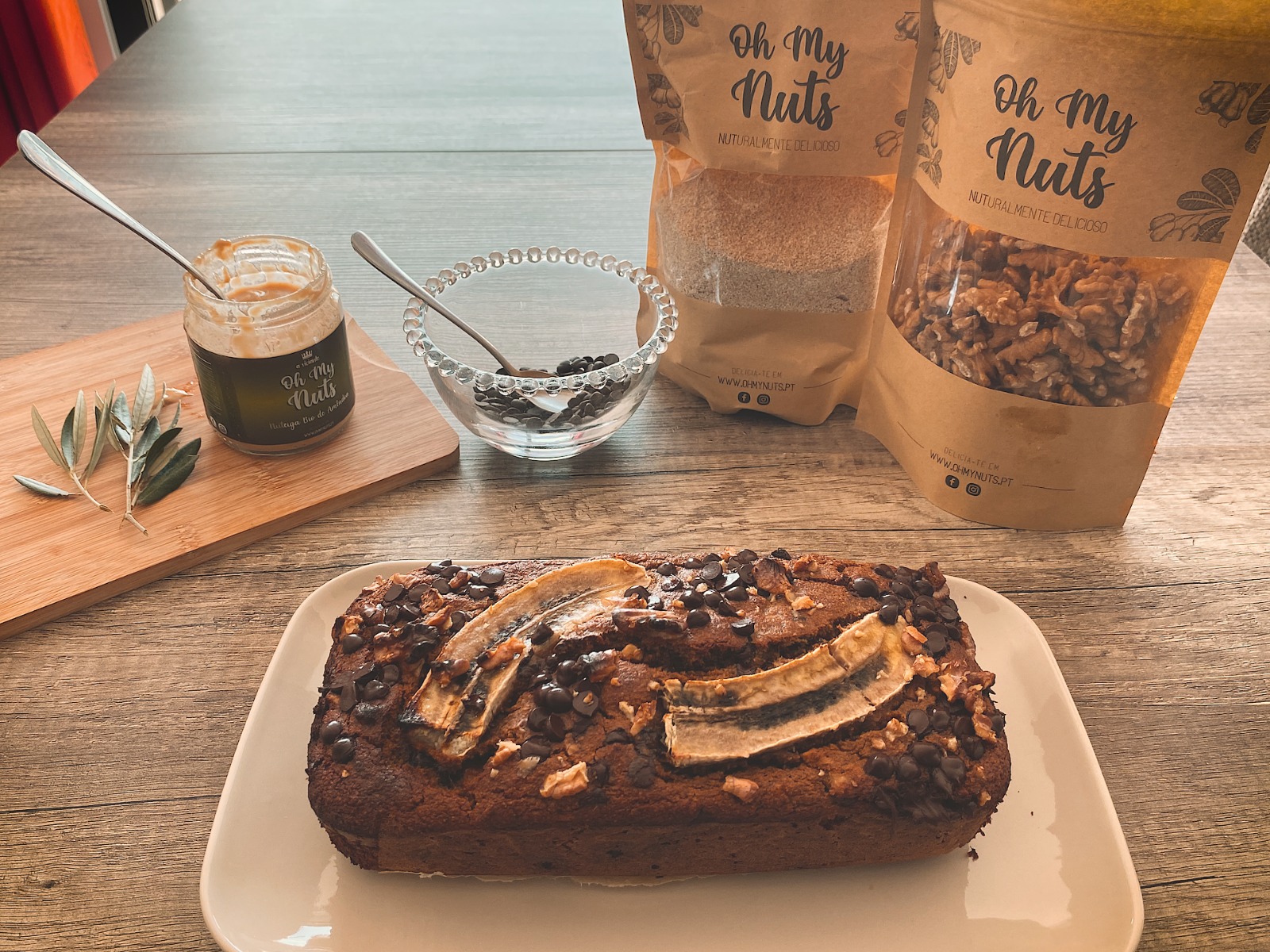 Bolo escuro com fatias de banana e pepitas de chocolate, frascos e pacotes de frutos secos Oh My Nuts numa mesa de madeira