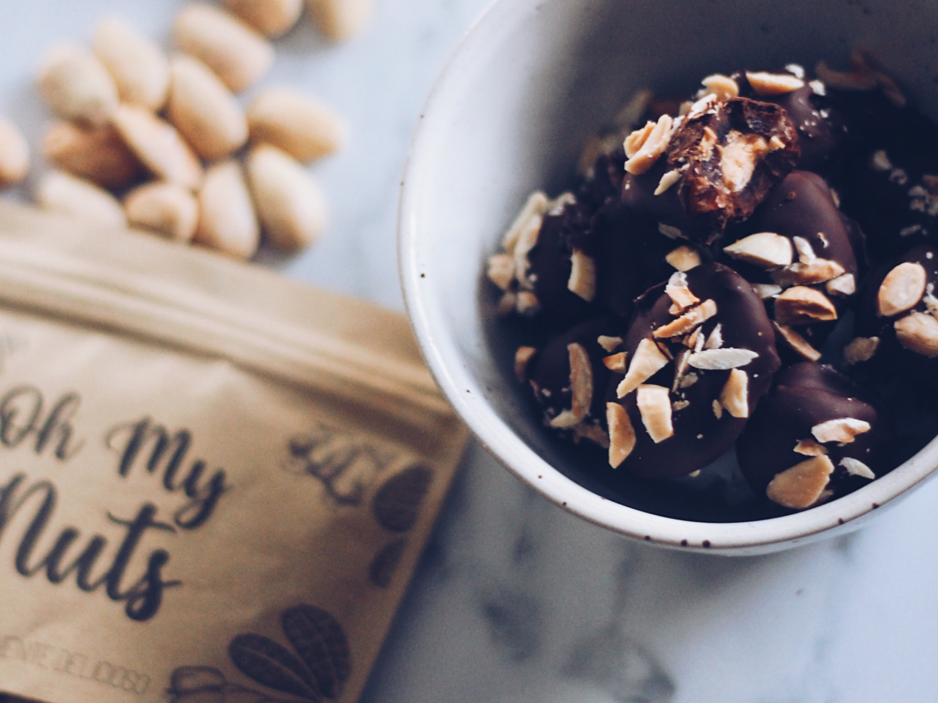Pacote de papel castanho e tigela branca com amendoins cobertos de chocolate e pedaços de amendoim