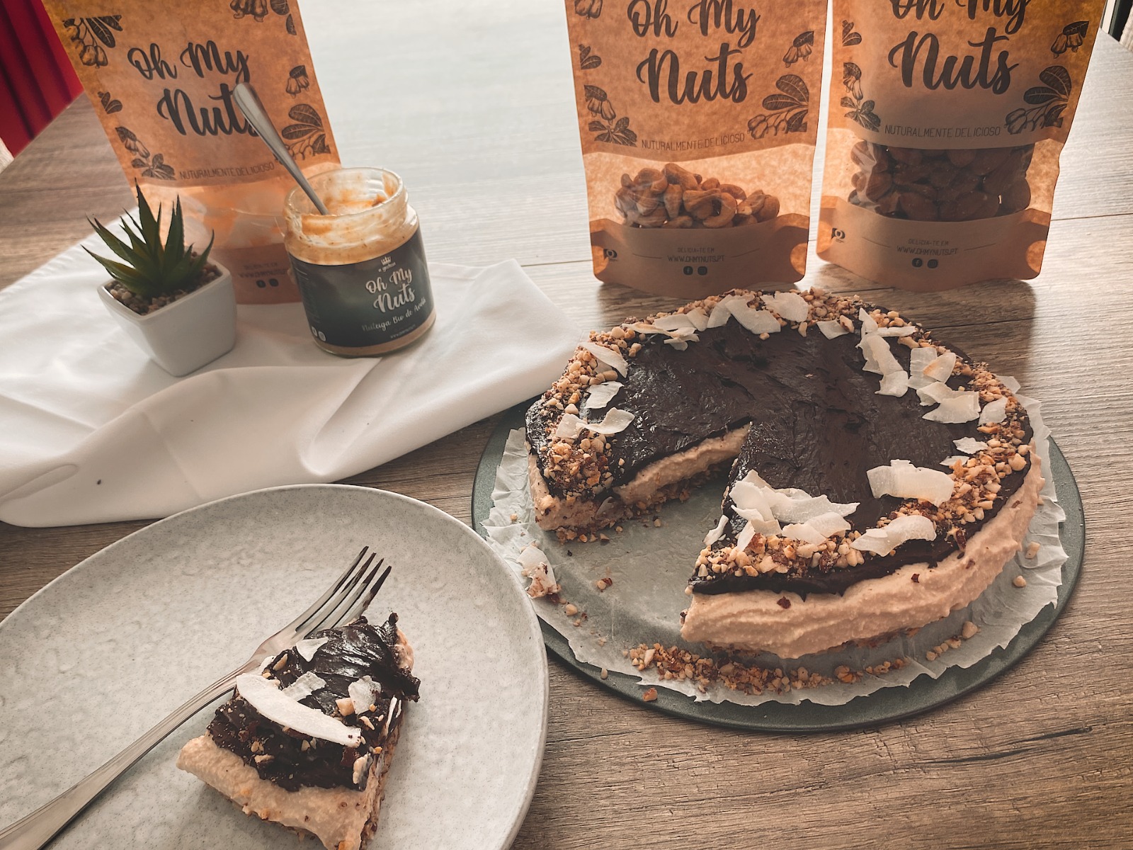 Bolo de chocolate com lascas de coco, frasco de compota e embalagens 'Oh My Nuts' numa mesa de madeira
