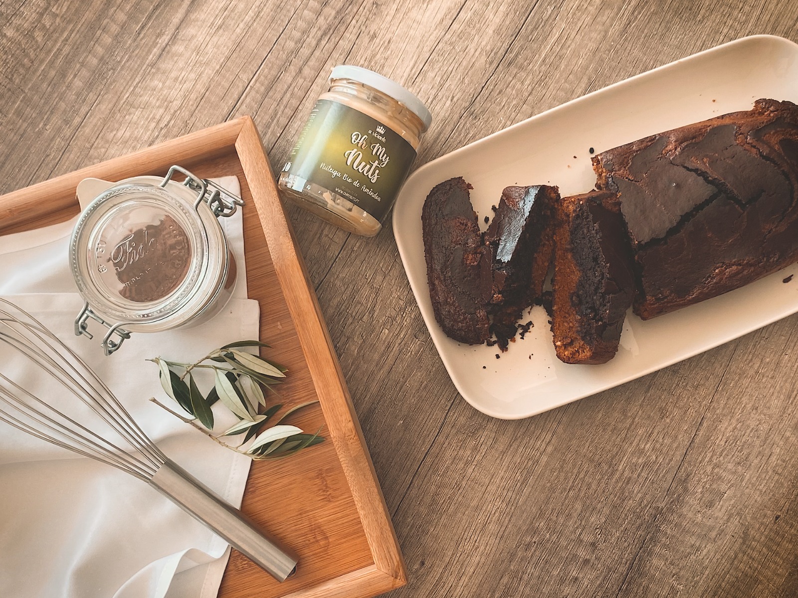 Bolo de chocolate cortado em prato, frascos com ingredientes, batedor e folhas numa bandeja de madeira.