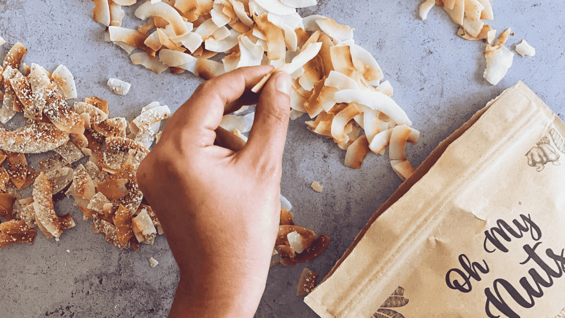 Mão a segurar lasca de coco torrado com embalagem castanha 'Oh My Nuts'