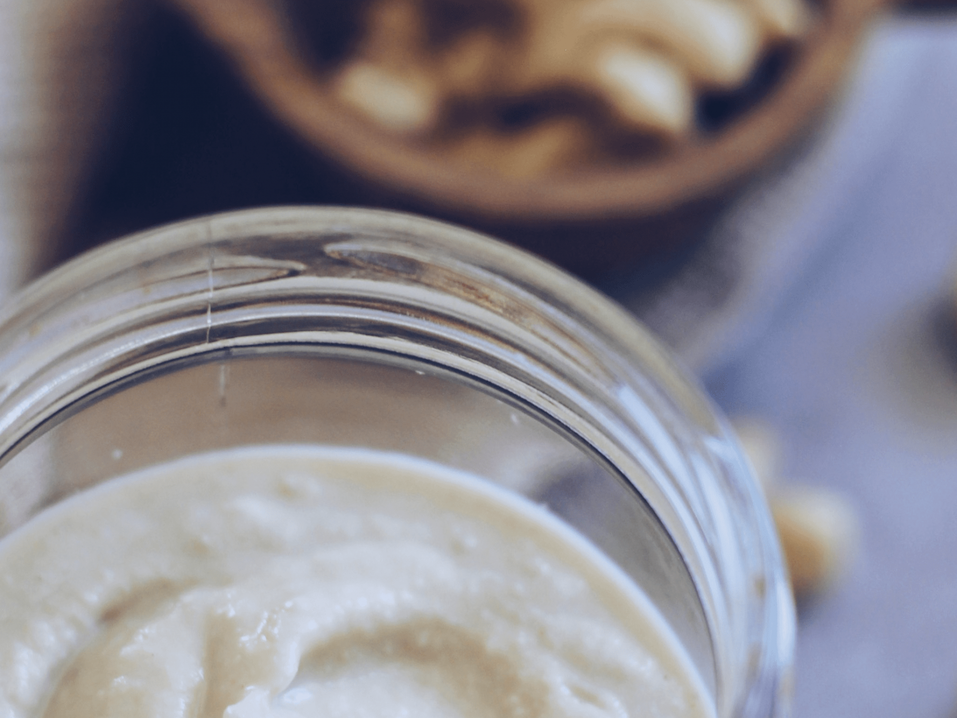 Frasco de vidro com pasta cremosa bege e recipiente de madeira com frutos secos ao fundo