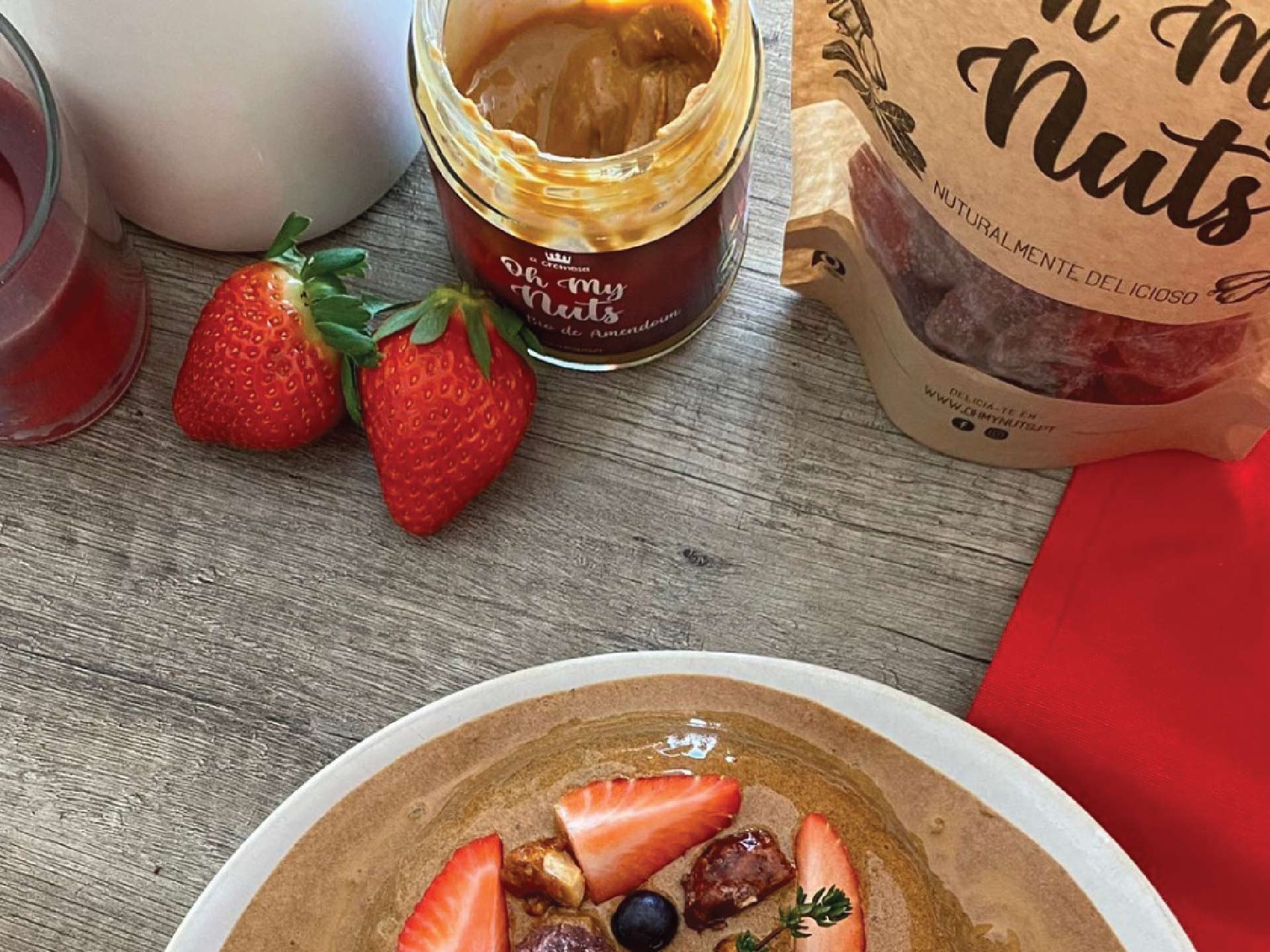 Frasco aberto de creme de amendoim, morangos, panquecas com fruta em prato, embalagem da marca 'do my nuts' e guardanapo vermelho sobre mesa de madeira.