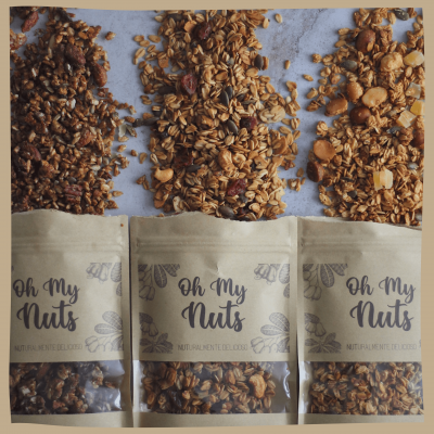 Três pacotes de granola 'Oh My Nuts' com produtos derramados sobre uma superfície cinza