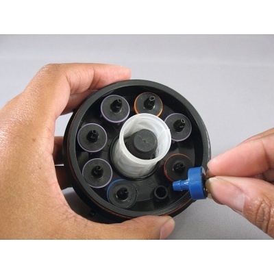 Mãos a segurar e inserir componente azul num suporte circular preto com cilindros coloridos