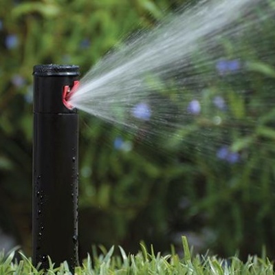 Aspersor preto a regar relva com plantas ao fundo