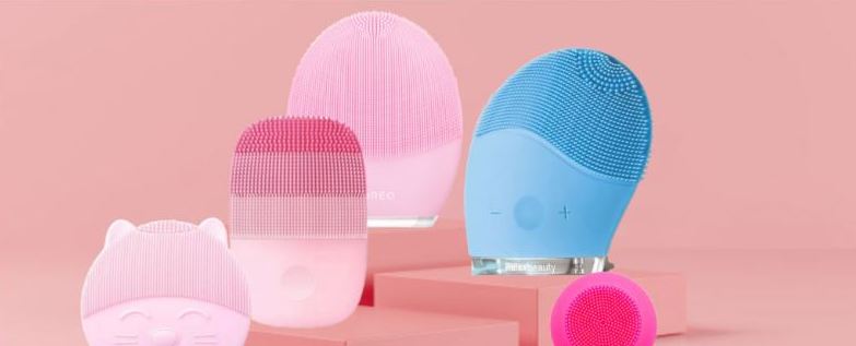 Dispositivos de limpeza facial em silicone coloridos com texturas de cerdas, sobre plataforma rosa com fundo rosa