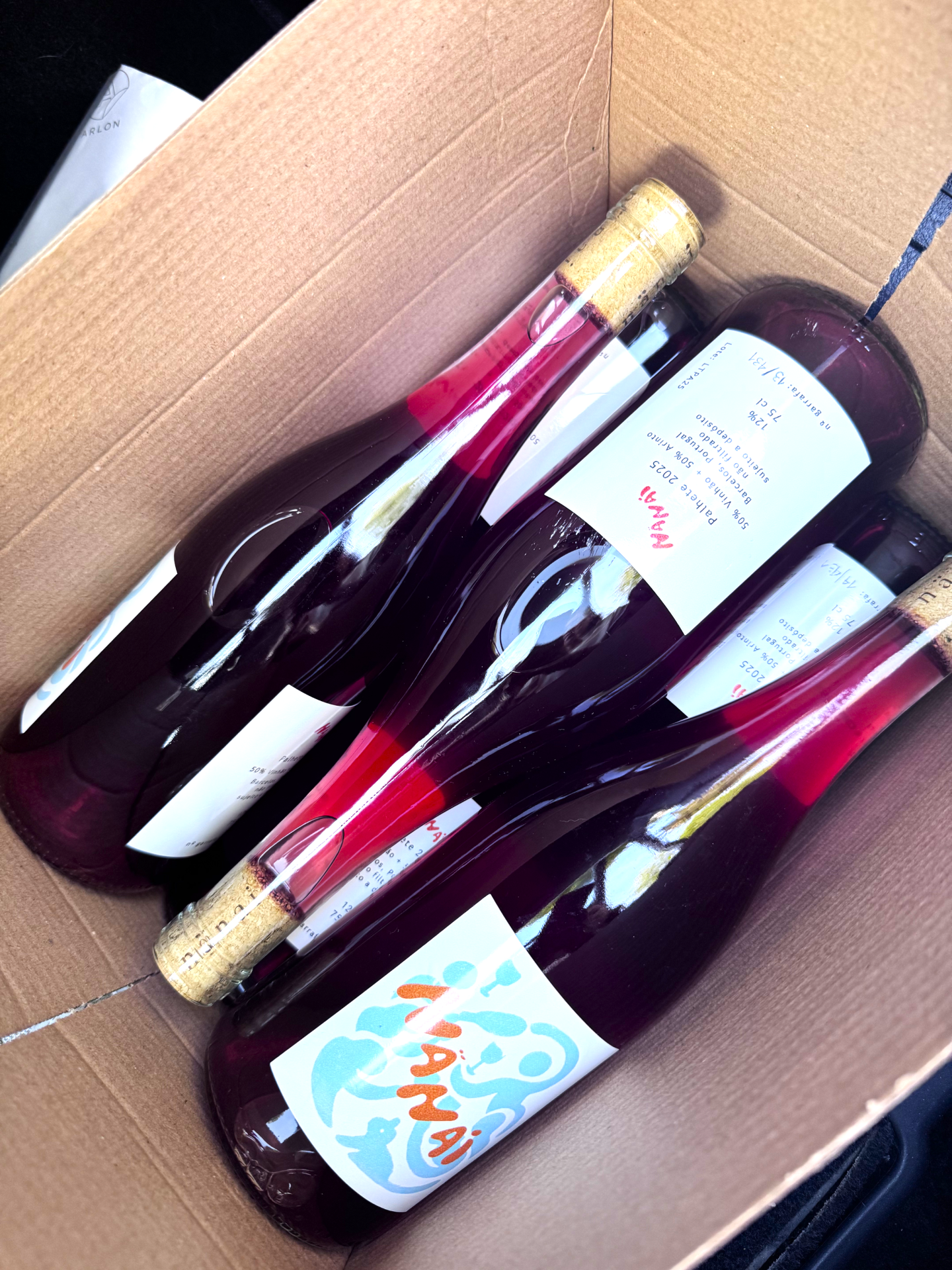 Garrafas de vinho em caixa de cartão com etiquetas brancas e texto em português.