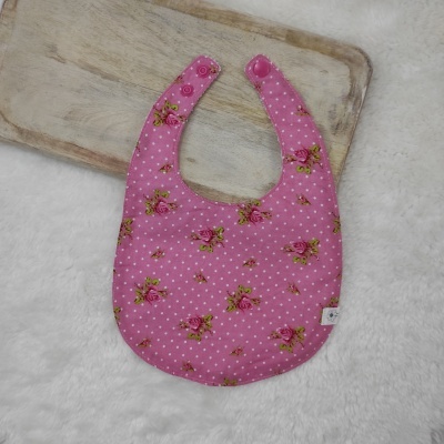 Babete rosa com padrão de rosas e bolinhas brancas sobre superfície branca e tábua de madeira