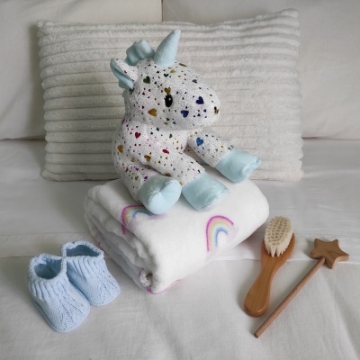 Conjunto de bebé com manta de arco-íris, brinquedo de unicórnio colorido, sapatinhos azuis, escova de cabelo e cabide de madeira sobre cama branca.