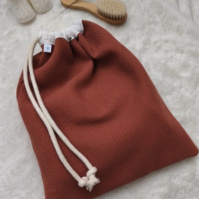 Saco de algodão castanho com cordão branco, escova de madeira com cerdas claras e botas de bebé brancas em malha sobre superfície felpuda branca.