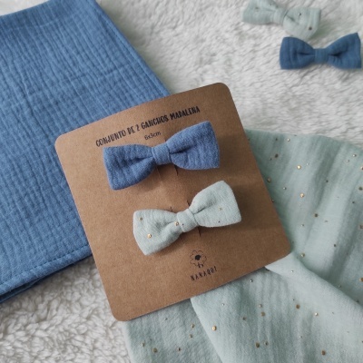 Conjunto de 2 ganchos laço azul e verde-água com bolinhas douradas, cartão castanho com texto e logo sobre tecido azul e superfície felpuda