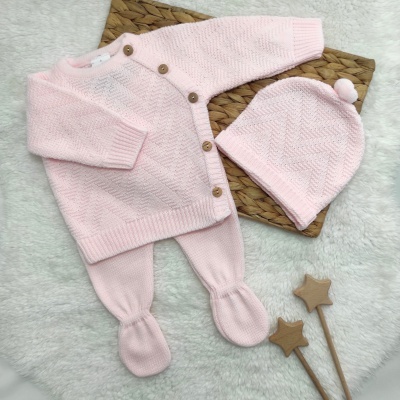 Conjunto de roupa de bebé rosa de malha com casaco, calças e gorro, sobre tapete branco com base castanha e dois pauzinhos com estrelas