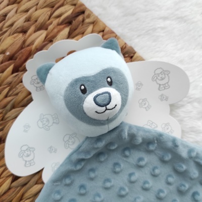 Pelúcia de urso cinzento e azul claro com tecido macio texturizado, sobre pano branco e tapete de fibras naturais