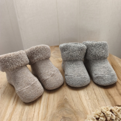 Dois pares de meias para bebé em tecido bege e cinzento claro com bainha felpuda sobre mesa de madeira clara.