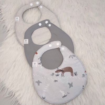 Três babetes de bebé em diferentes tons de cinza com diferentes padrões, apoiados sobre um tecido branco peludo.