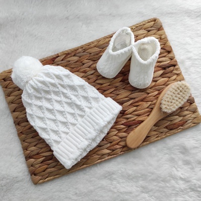 Gorro branco de malha, sapatilhas brancas felpudas e escova de madeira em base entrelaçada castanha sobre fundo branco.