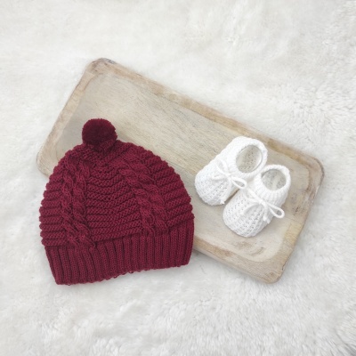 Gorro borgonha tricotado com pompom e sapatinhos brancos tricotados sobre tábua de madeira