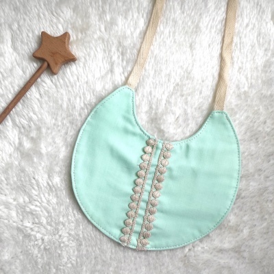 Babete de bebé verde água com bordado bege e fitas de algodão cru sobre fundo branco felpudo