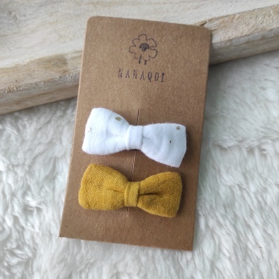 Dois laços de tecido branco e mostarda fixados num cartão castanho com logótipo MANAAQI