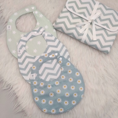 Três babetes coloridos com padrões de estrelas, zig-zag e margaridas sobre superfície felpuda branca, junto com toalha dobrada com laço.