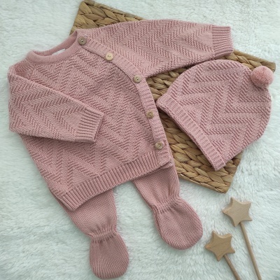 Conjunto de roupa de bebé rosa em malha com casaco, calças e gorro com pompom sobre tapete trançado e fundo branco