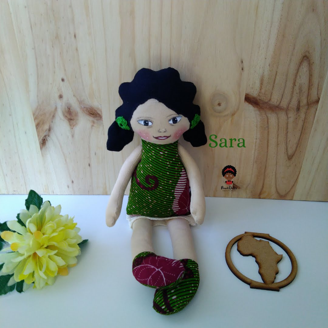 Sara Boneca de pano artesanal com vestido verde e vermelho, sentada com flor e detalhe de madeira ao lado