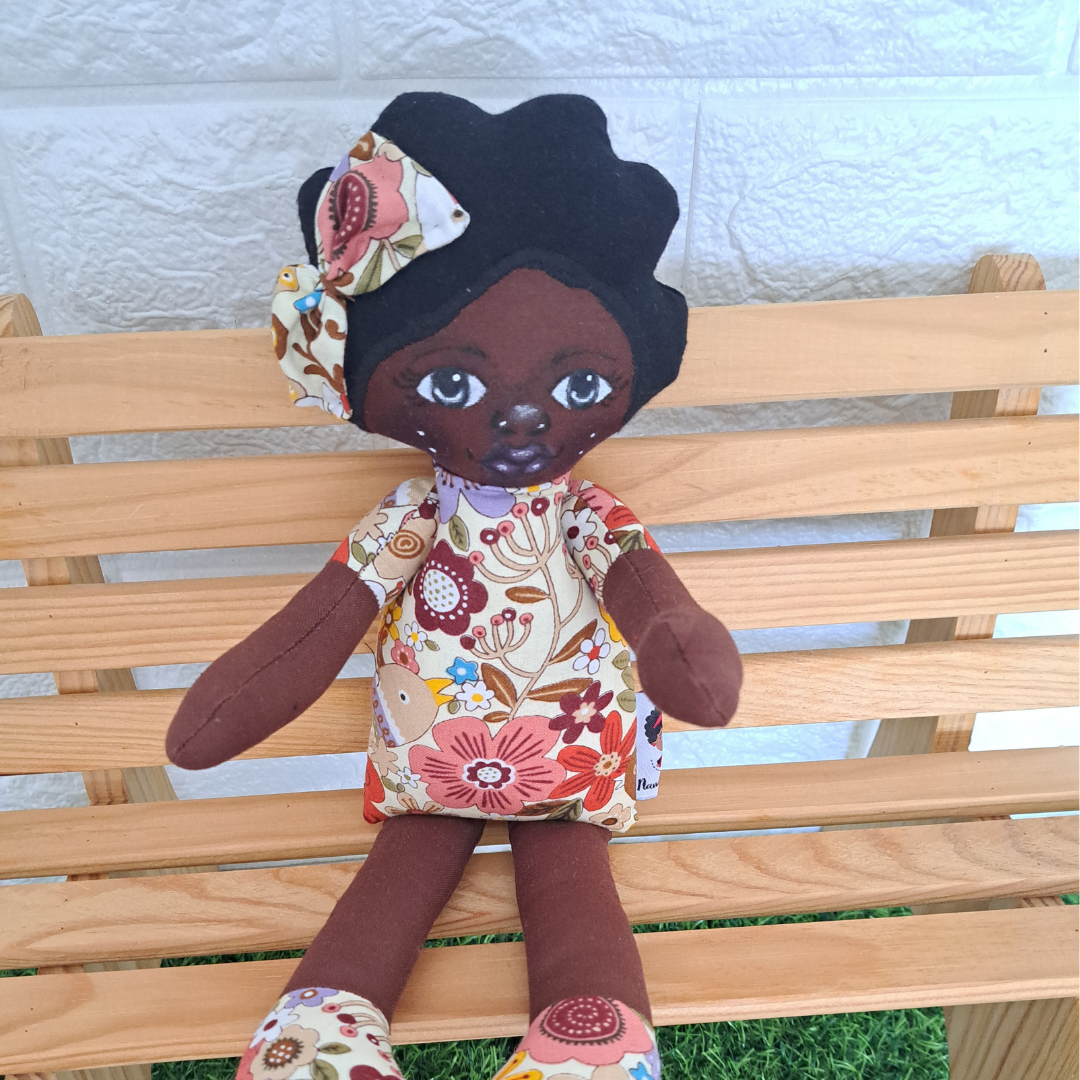 Keva Boneca de tecido castanha com vestido floral sentada num banco de madeira