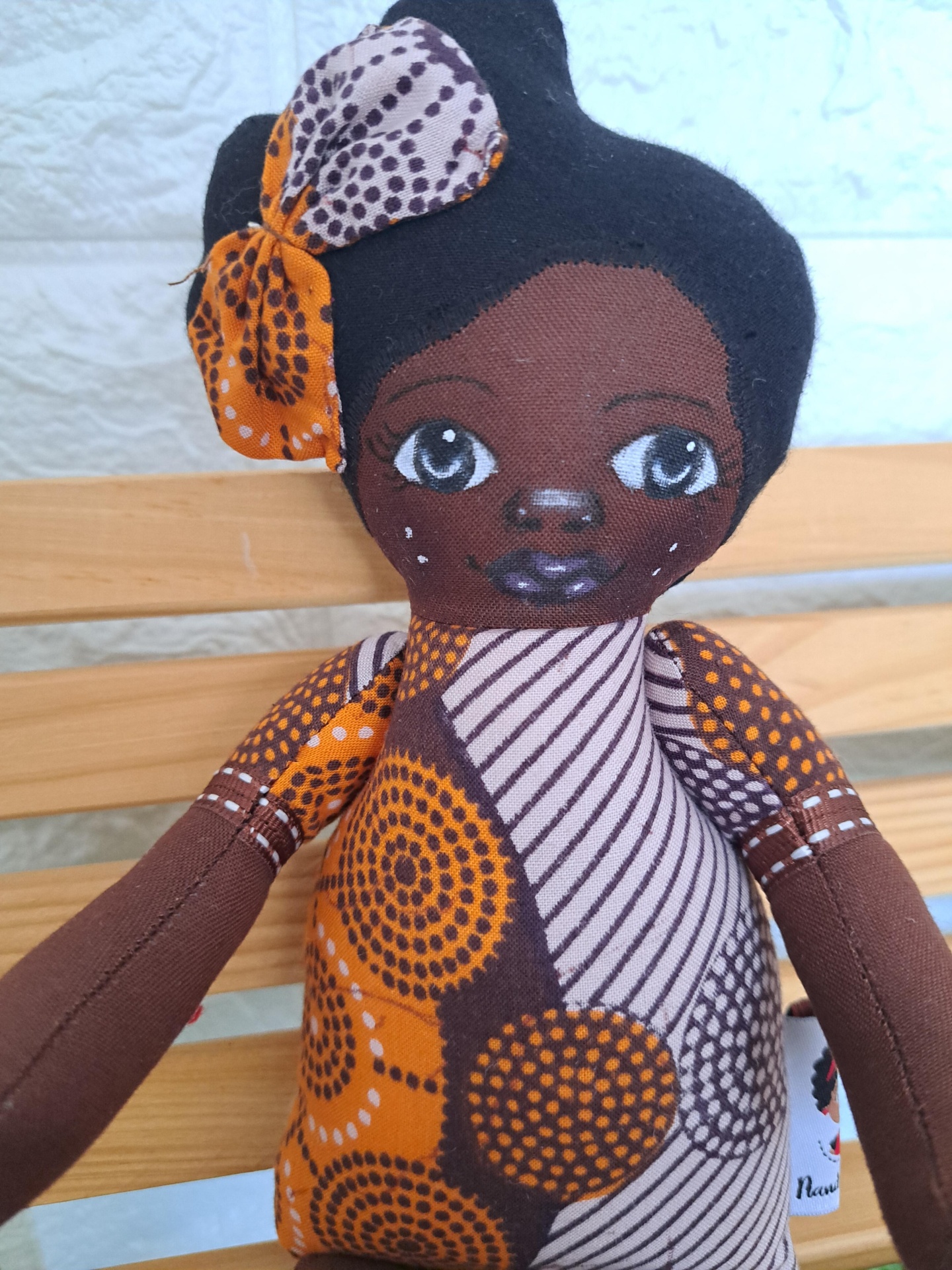 Sona Boneca de tecido com padrão laranja e castanho, cabelo preto e laço, sentada em banco de madeira.