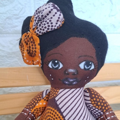 Boneca de pano com rosto pintado, cabelo preto e laço laranja e púrpura.