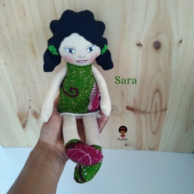 Boneca de pano com vestido verde e rosa segurada diante de fundo de madeira clara com nome Sara