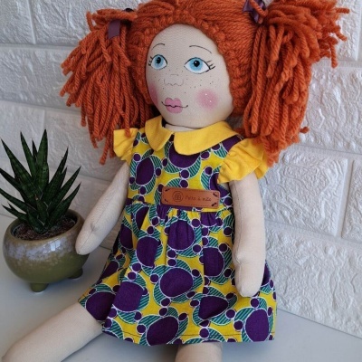 Boneca de tecido com vestido amarelo e padrão roxo sentada junto a planta em vaso