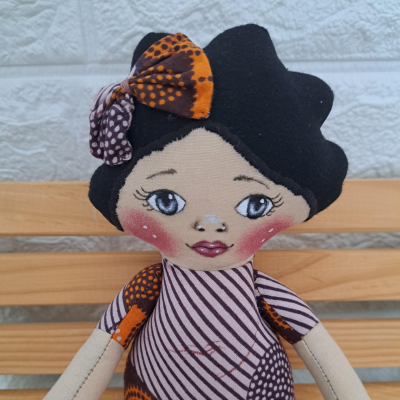 Boneca de tecido com padrão colorido e arco na cabeça sentada num banco de madeira