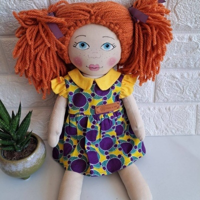 Boneca de tecido com vestido colorido e cabelo laranja de lã sentada ao lado de planta em vaso
