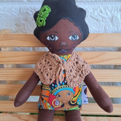 Boneca de pano colorida com vestido étnico e capa de crochê sentada em banco de madeira