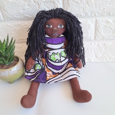 Boneca de pano com cabelo de lã e vestido colorido ao lado de planta num vaso