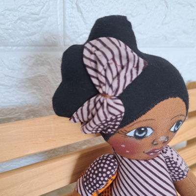 Boneca de pano com vestido listrado roxo e branco, sentada num banco de madeira