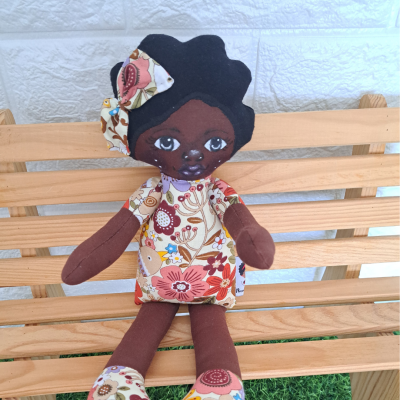 Boneca de tecido castanha com vestido floral sentada num banco de madeira