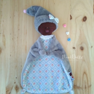 Boneca de tecido com roupa azul e cinzenta, com cabeça castanha e gorro com pompom, em superfície de madeira clara.