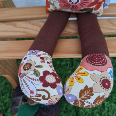 Pernas de boneca com tecido castanho escuro e pés florais coloridos sentada num banco de madeira