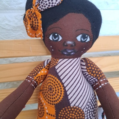 Boneca de tecido com padrão laranja e castanho, cabelo preto e laço, sentada em banco de madeira.