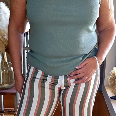 Mulher a usar top verde água sem mangas e calças listradas verticais em tons de verde, branco e rosa, pulseira de pérolas na mão direita, interior com móveis e plantas.