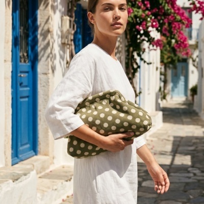 Mulher com vestido branco segura clutch verde de bolinhas em rua com portas azuis e flores.