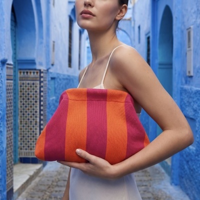 Mulher com vestido branco segurando bolsa listrada rosa e laranja em rua azul