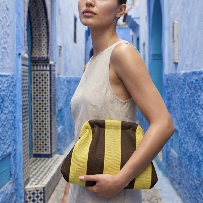 Mulher com vestido bege a segurar bolsa riscada amarela e castanha numa rua azul