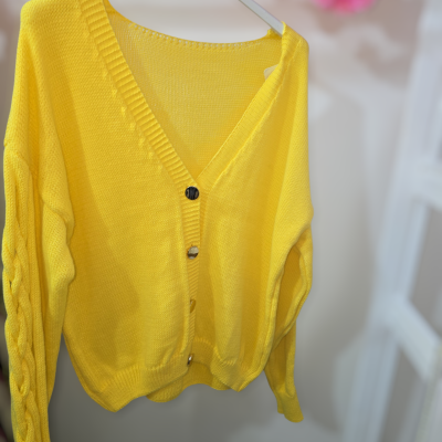 Cardigã amarelo em malha com botões metálicos num cabide branco.